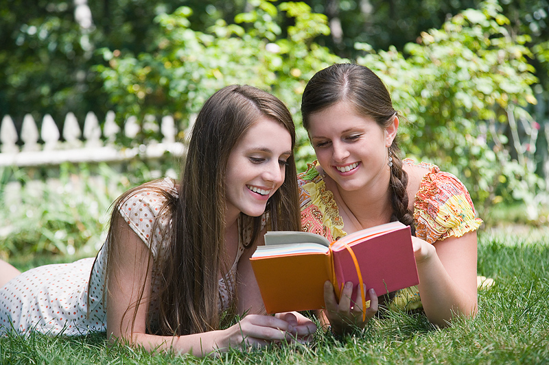 Två flickor ligger på gräset och läser i samma bok